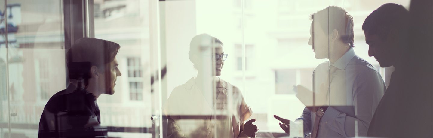 Quatre personnes discutent debout dans un bureau. Cliché pris derrière une vitre